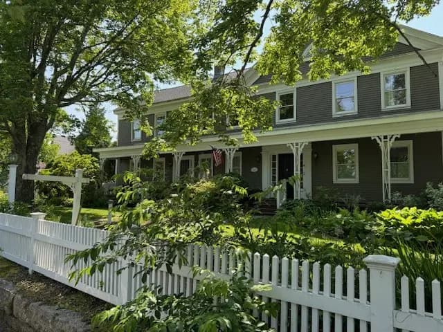 A charming two-story gray house with a white picket fence and lush greenery surrounding it.