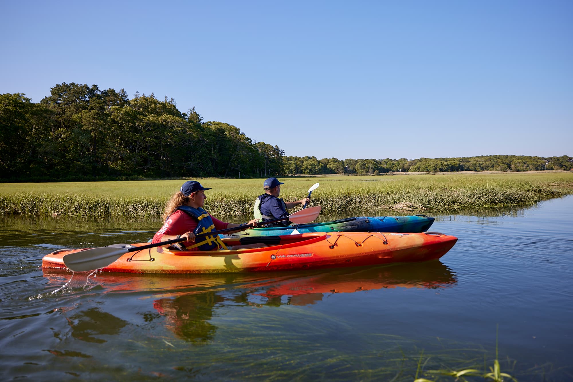 Two people paddle kayaks through a calm, grassy waterway under a clear blue sky.