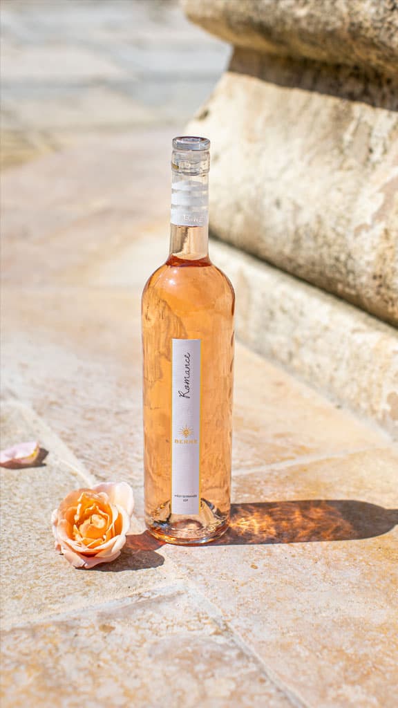 A bottle of rosé wine next to a delicate peach rose on a sunlit stone surface.