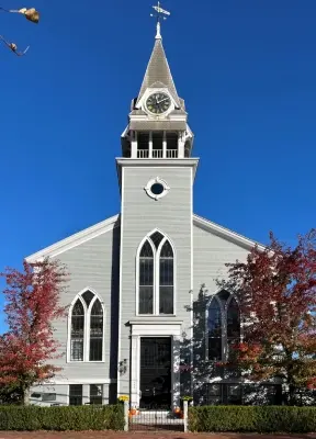 A gray church with a clock tower and arched windows surrounded by autumn trees against a clear blue sky.