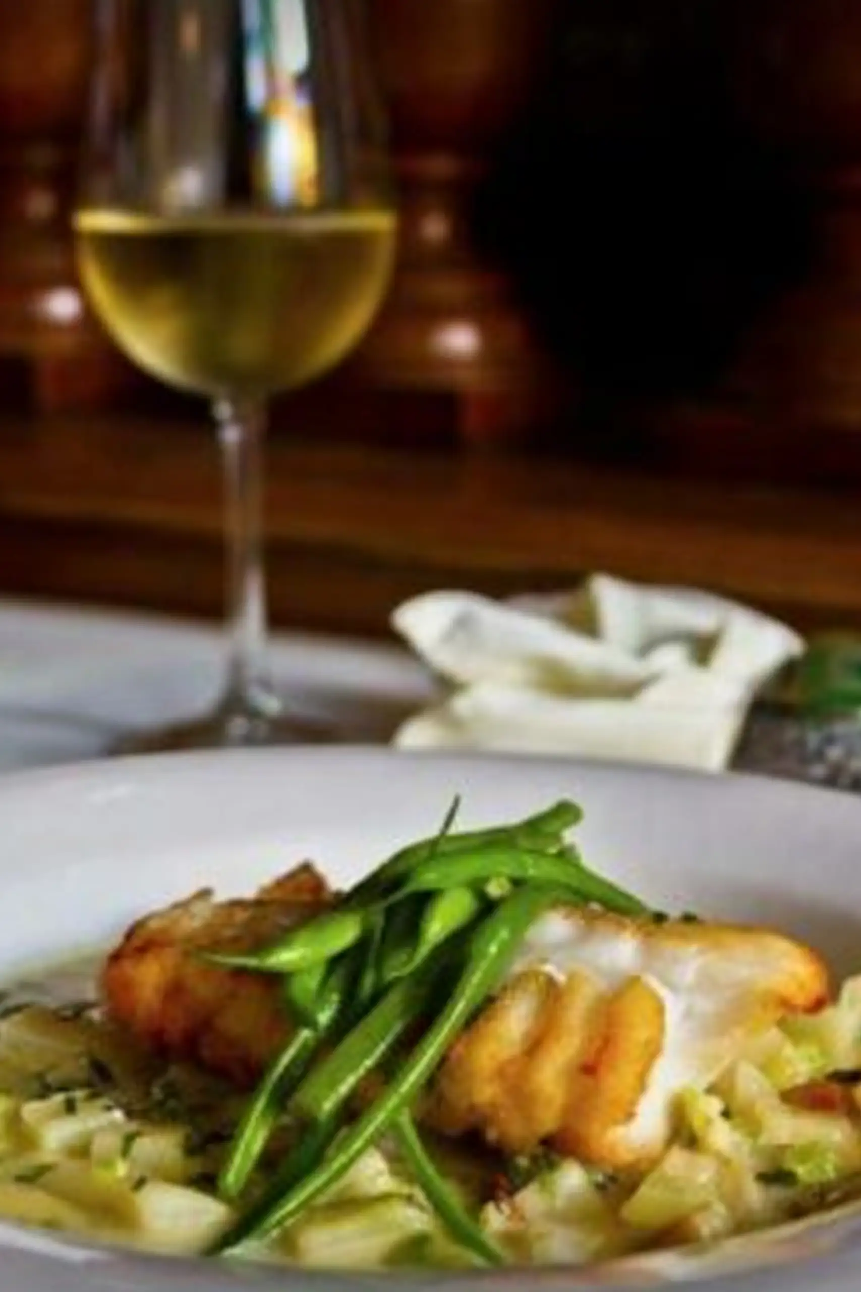A plate of fish topped with green beans, served over pasta with a glass of white wine in the background.