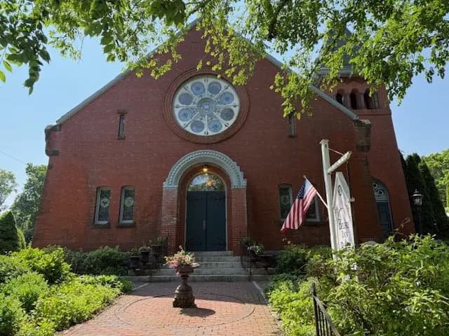 Red brick church with a circular stained glass window and an American flag in front, surrounded by greenery.